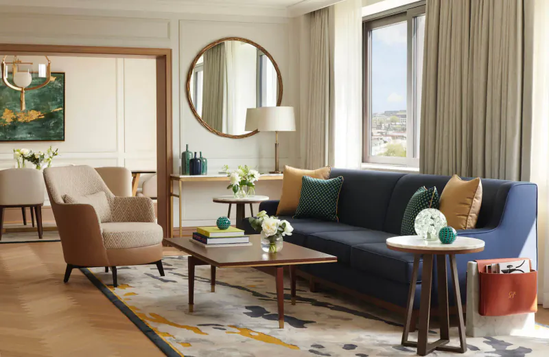 Elegant luxury hotel suite with navy sofa, armchairs, coffee tables, flowers, and city view.