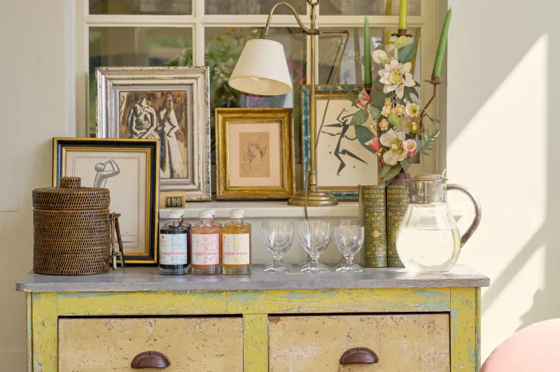 Vintage yellow dresser with Christopher’s Cordials bottles, antique frames, lamp, flowers, and glassware in sunlit gallery window.