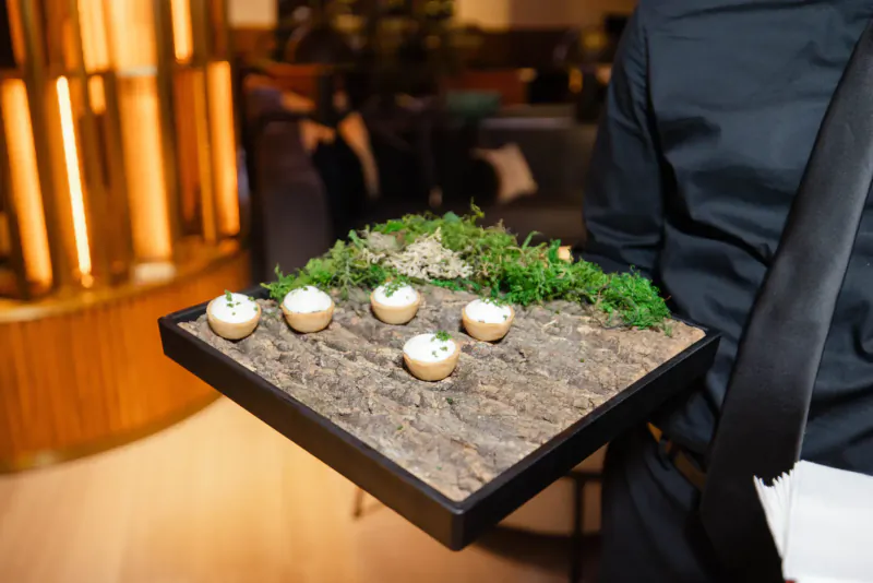 Chef holding tray of white-topped appetizers on mossy surface in elegant venue