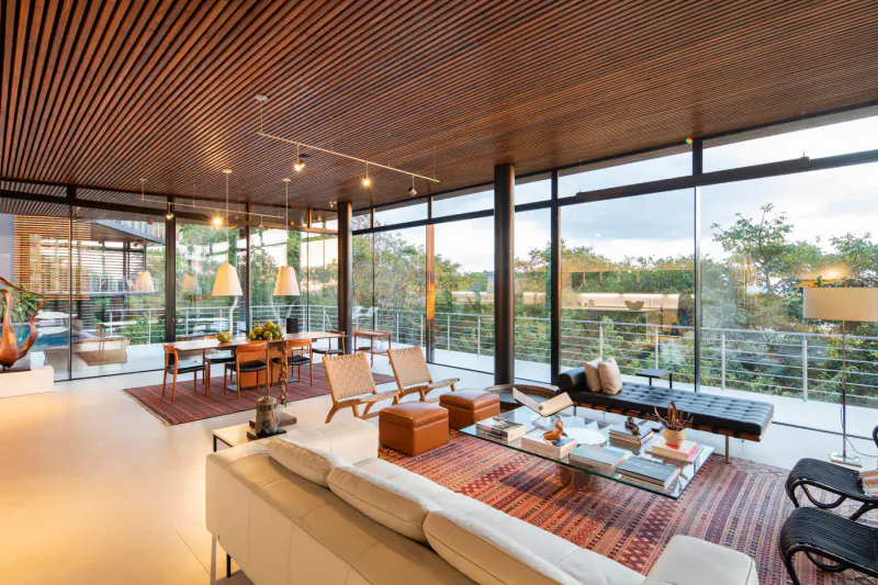 Modern luxury living room with wooden slat ceiling, white sofa, dining table, large windows to lush garden balcony.