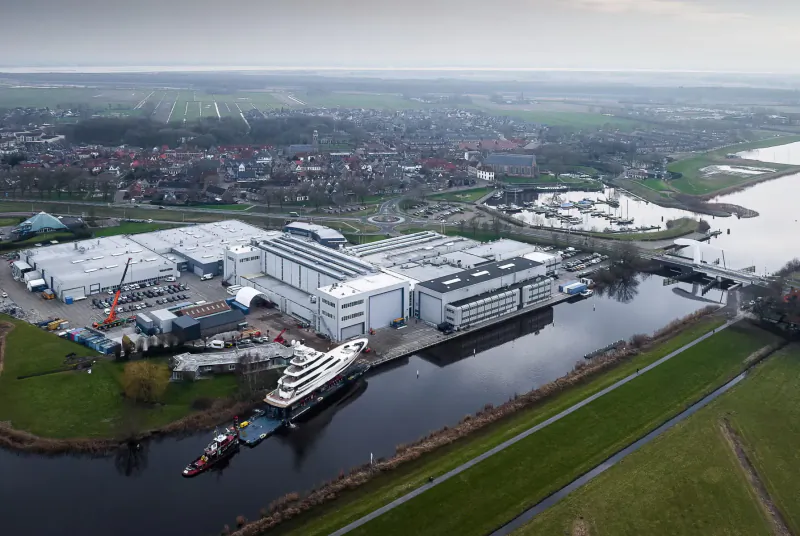 Aerial view of white yacht shipyard with docked superyacht, warehouses, cranes, canal, fields, and village.
