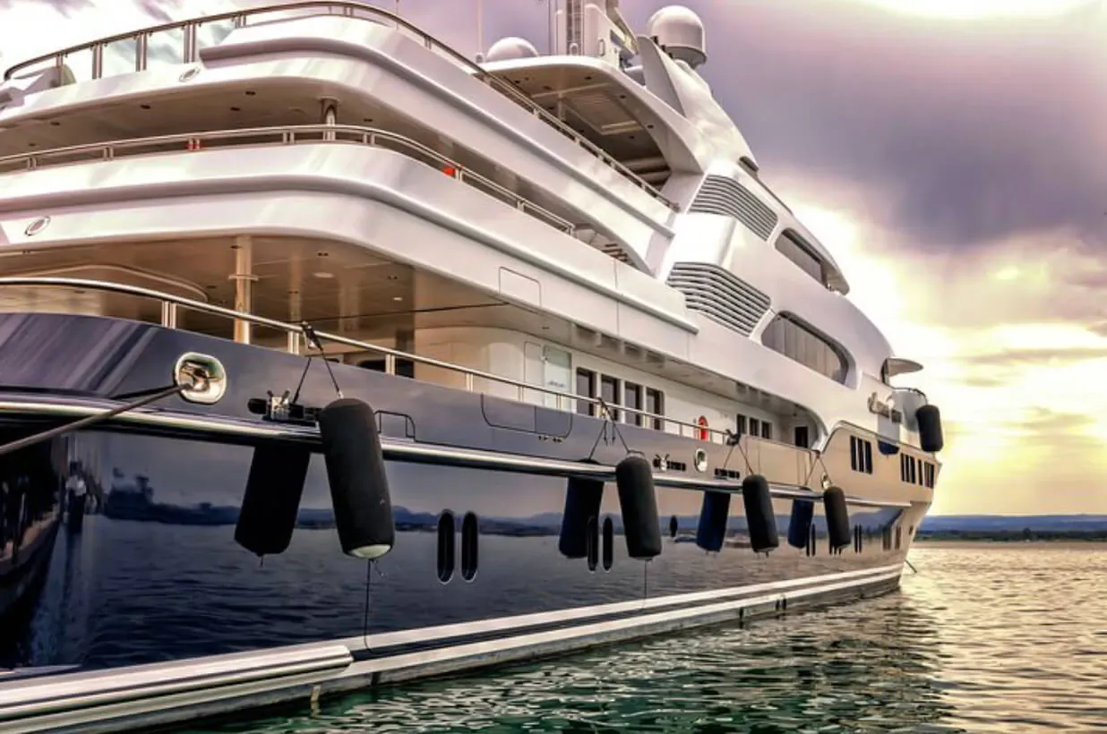 Luxurious white superyacht docked at marina with fenders, calm water, sunset sky.