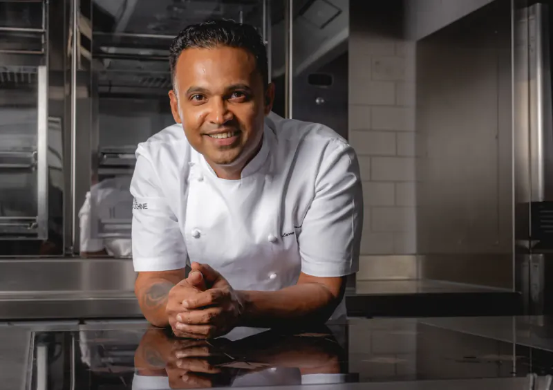 Smiling male chef in white uniform, leaning on kitchen counter with clasped hands amid stainless steel equipment.