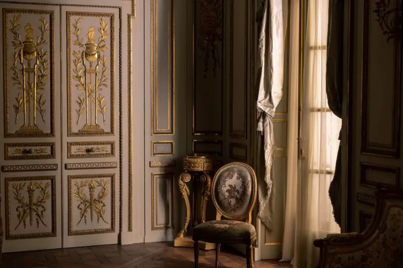 Elegant Regency-style interior with ornate gilded doors, floral armchair, pedestal table, and sheer curtains by window.