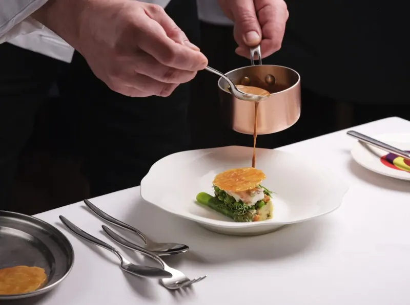 Chef in white uniform pours sauce from copper pitcher onto plated dish with greens and crab in cooking masterclass