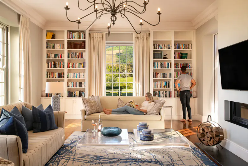 Elegant library lounge at Brookdale Wine Estate with woman relaxing on sofa, another browsing bookshelves, garden view.