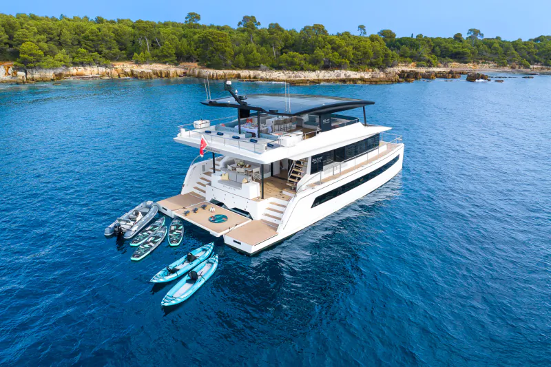 Aerial view of luxury white Silent Grace catamaran yacht with kayaks, docked in turquoise waters near green coastal cliffs.