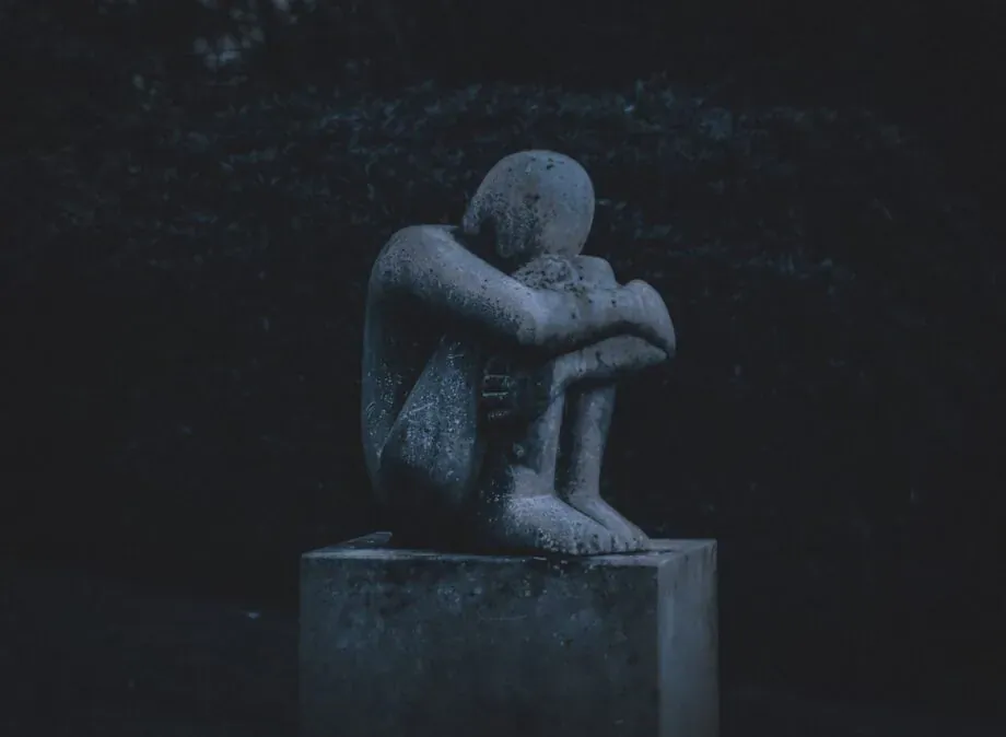 Stone statue of a nude figure crouched with head in hands on pedestal, evoking depression amid dark foliage.