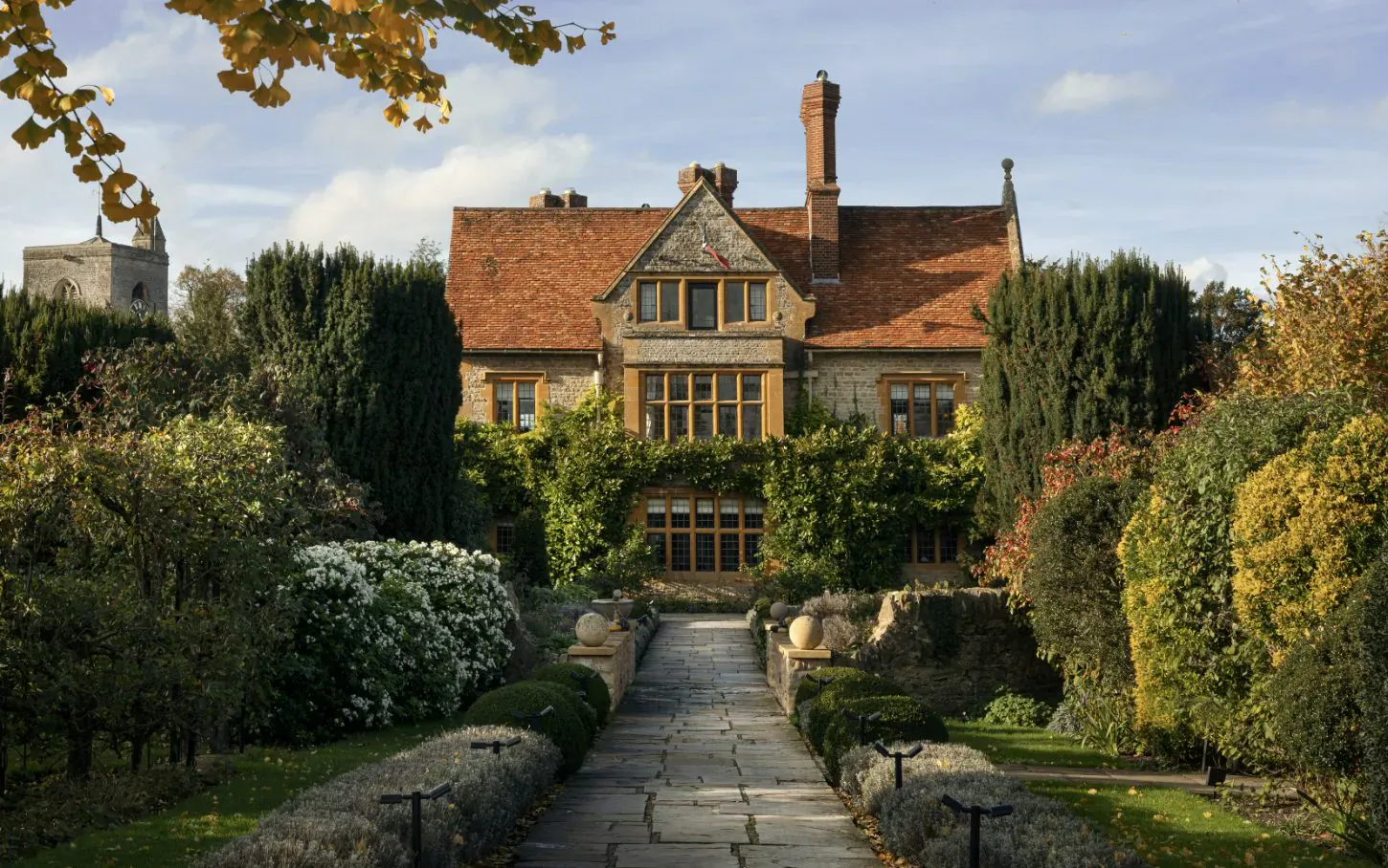 Le Manoir aux Quat'Saisons manor house with red-tiled roof, gardens, stone path, and autumn foliage under blue sky