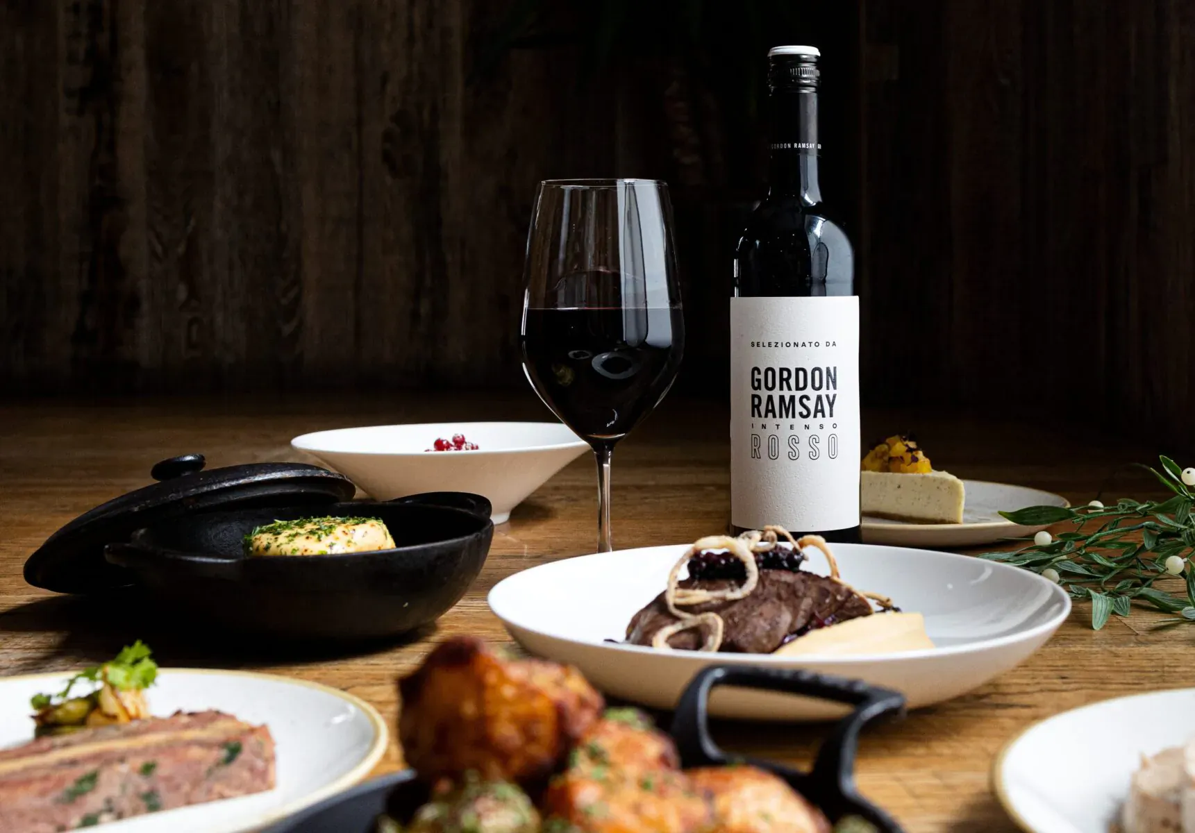 Gordon Ramsay wine bottle beside red wine glass, steak, mushrooms, cheese on wooden table with Italian dishes