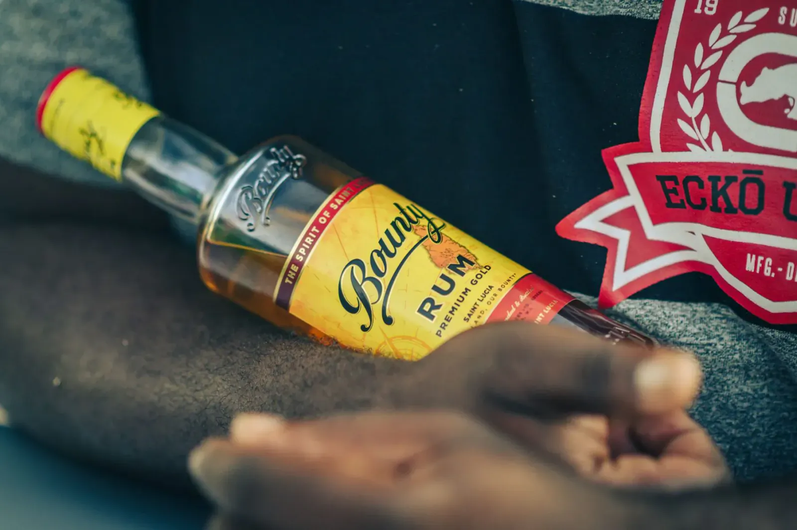 Bounty Rum bottle held in dark hand on black clothing with red Ecko Unltd patch, Bounty Rum label visible.