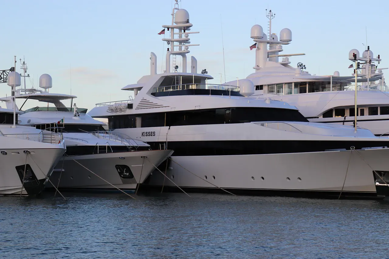 Several white luxury yachts docked at marina, sleek designs with antennas and flags, calm water backdrop.