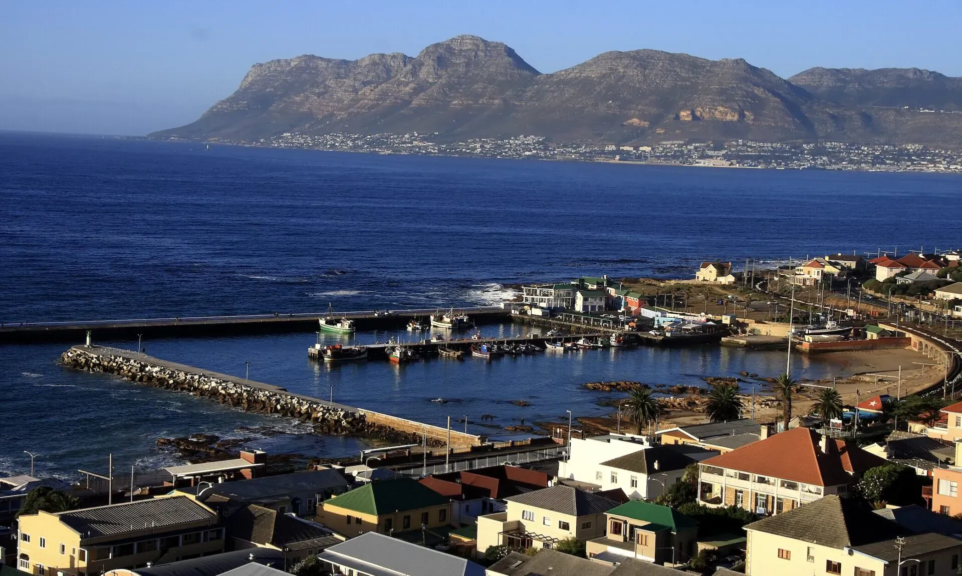Aerial view of Kalk Bay harbour in Cape Town, with fishing boats, colorful houses, and Table Mountain range across the ocean.