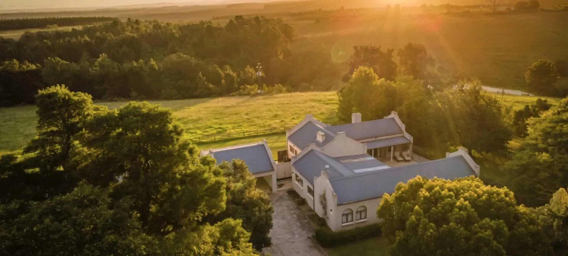 Aerial view of Kay and Monty Vineyard Boutique Wine Estate in Plettenberg, South Africa, at golden hour sunset amid green fields and trees.