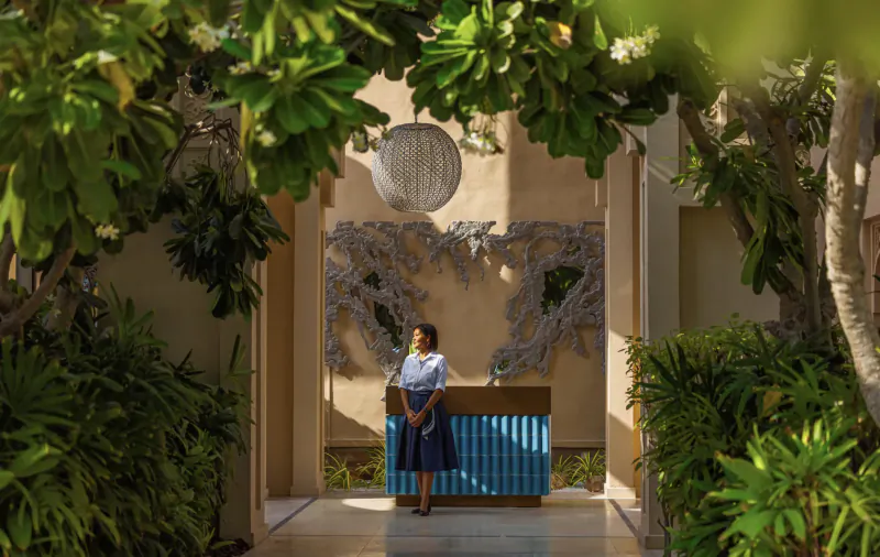 Woman in white blouse and blue skirt stands at blue reception desk in lush tropical Sea Fu resort entrance with greenery and lantern