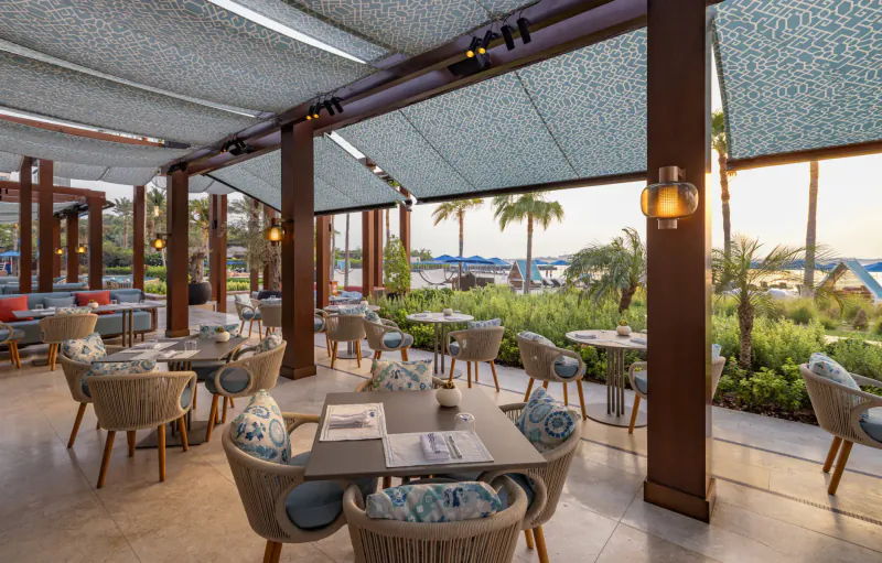 Elegant outdoor dining area at Sea Fu, Four Seasons Resort Dubai, with rattan tables, blue cushions, palms, and beach sunset view.