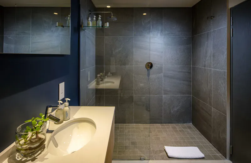 Modern hotel bathroom with glass shower, vanity sink, plants, white towel on tile floor.