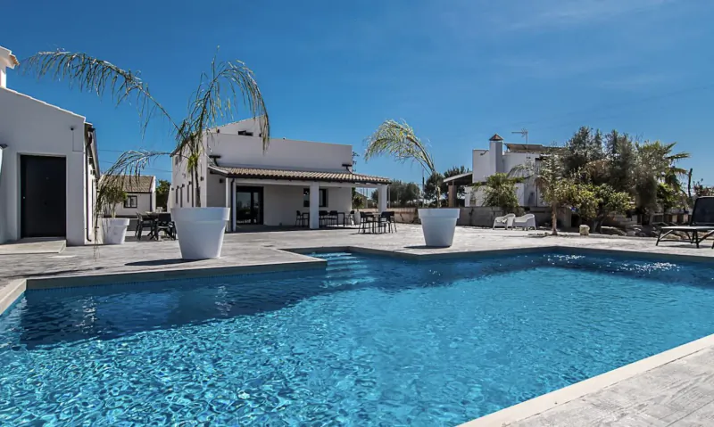 White modern villas with blue infinity pool, lounge chairs, large white pots, and palm trees under clear blue sky at La Vuelta Boutique Hotel.