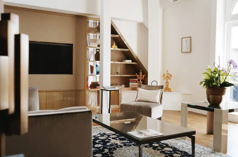 Modern luxury living room with beige sofas, glass coffee table, TV, bookshelves, plants, and gold statue.