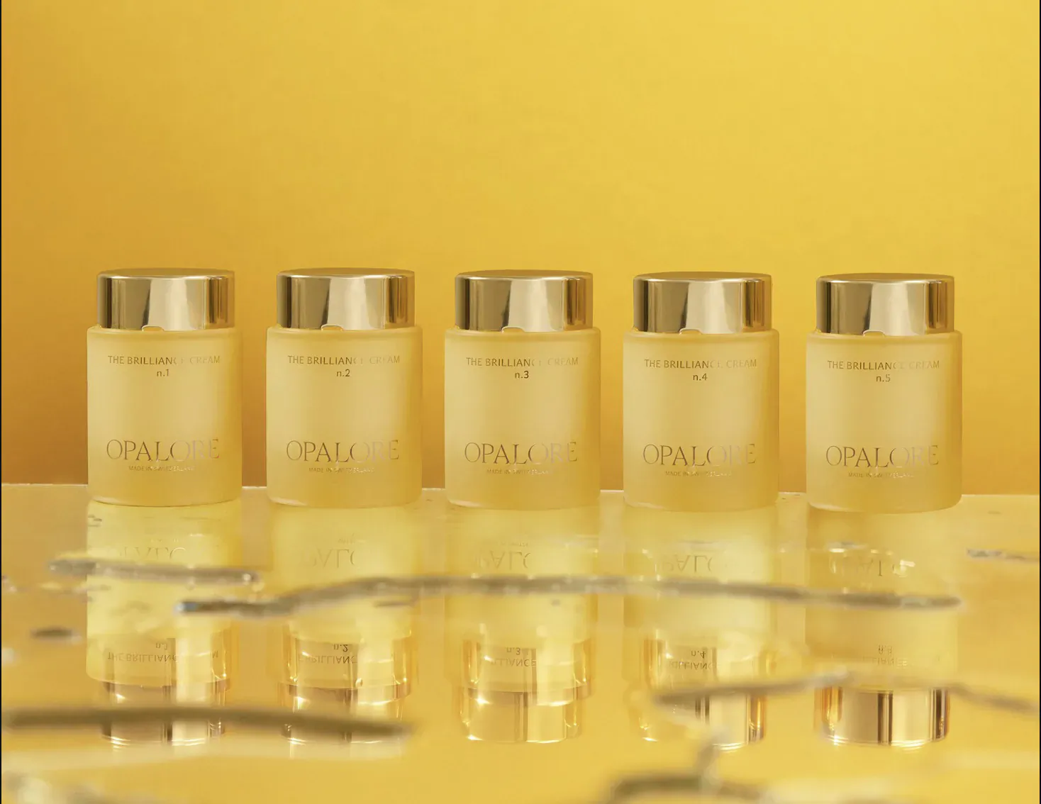 Five gold-capped Opalore skincare bottles in a row on reflective yellow surface, 'The Brilliance of skin' featured.
