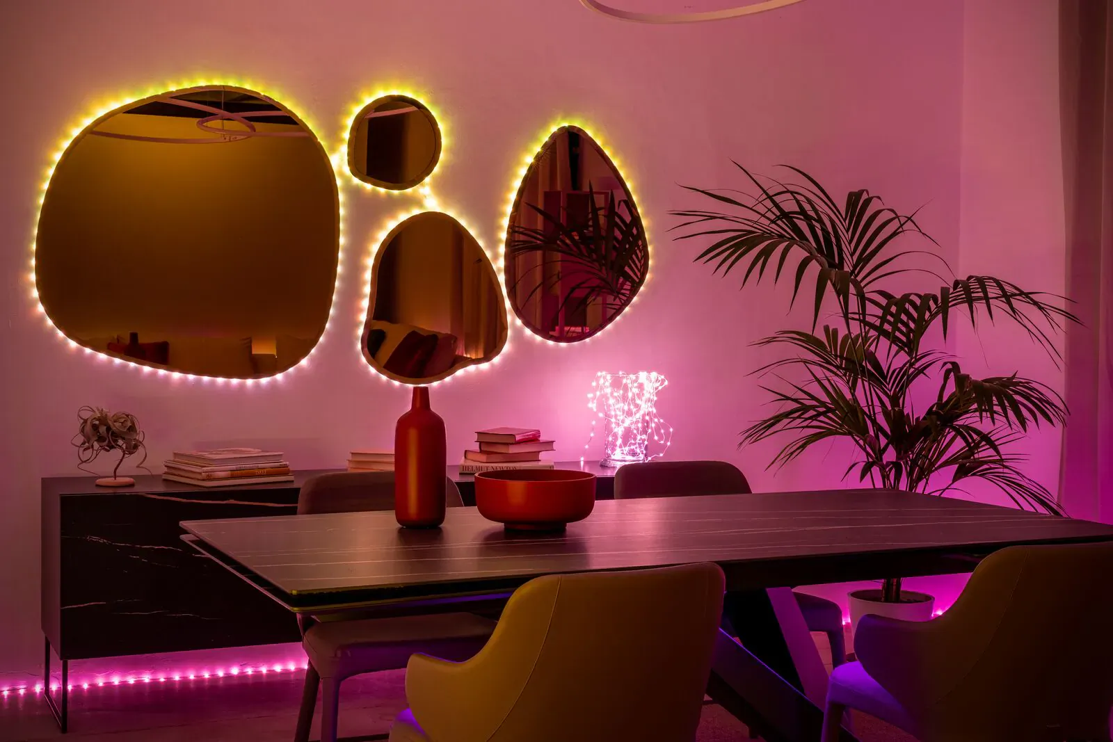 Modern dining room with wooden table, yellow LED-lit abstract mirrors, pink ambient lights, red vase, plants, and chairs.