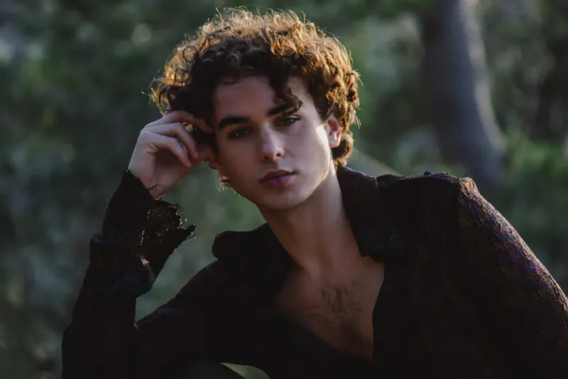 Javi Costa Polo with curly hair, hand on chin, thoughtful pose in black shirt against green trees