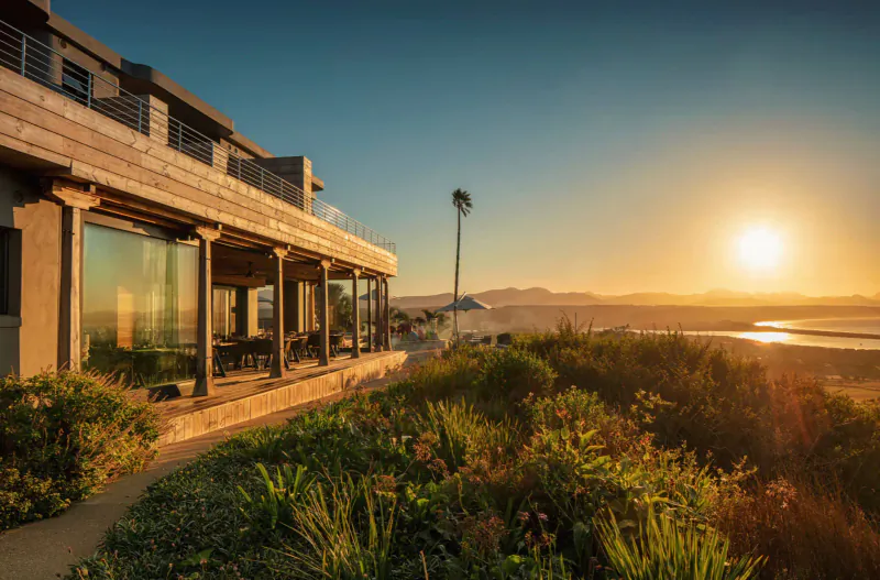 Sky Villa Boutique Hotel on coastal cliff at sunset, with ocean views, modern wooden exterior, balcony, and lush greenery.