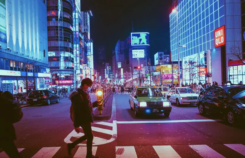 Man with backpack crossing neon-lit Tokyo street at night at crosswalk amid traffic and UNIQLO signs.