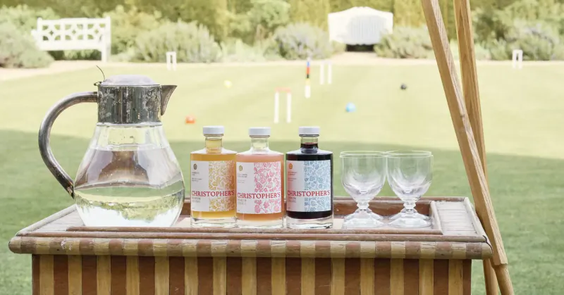 Outdoor croquet match with tray of water pitcher, colorful Christopher’s Cordials bottles, and glasses on grass.
