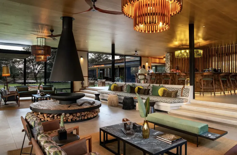 Spacious lodge interior at Tanda Tula with central black chimney fireplace, wooden surrounds, orange chandelier, green cushions, bar, and large windows.