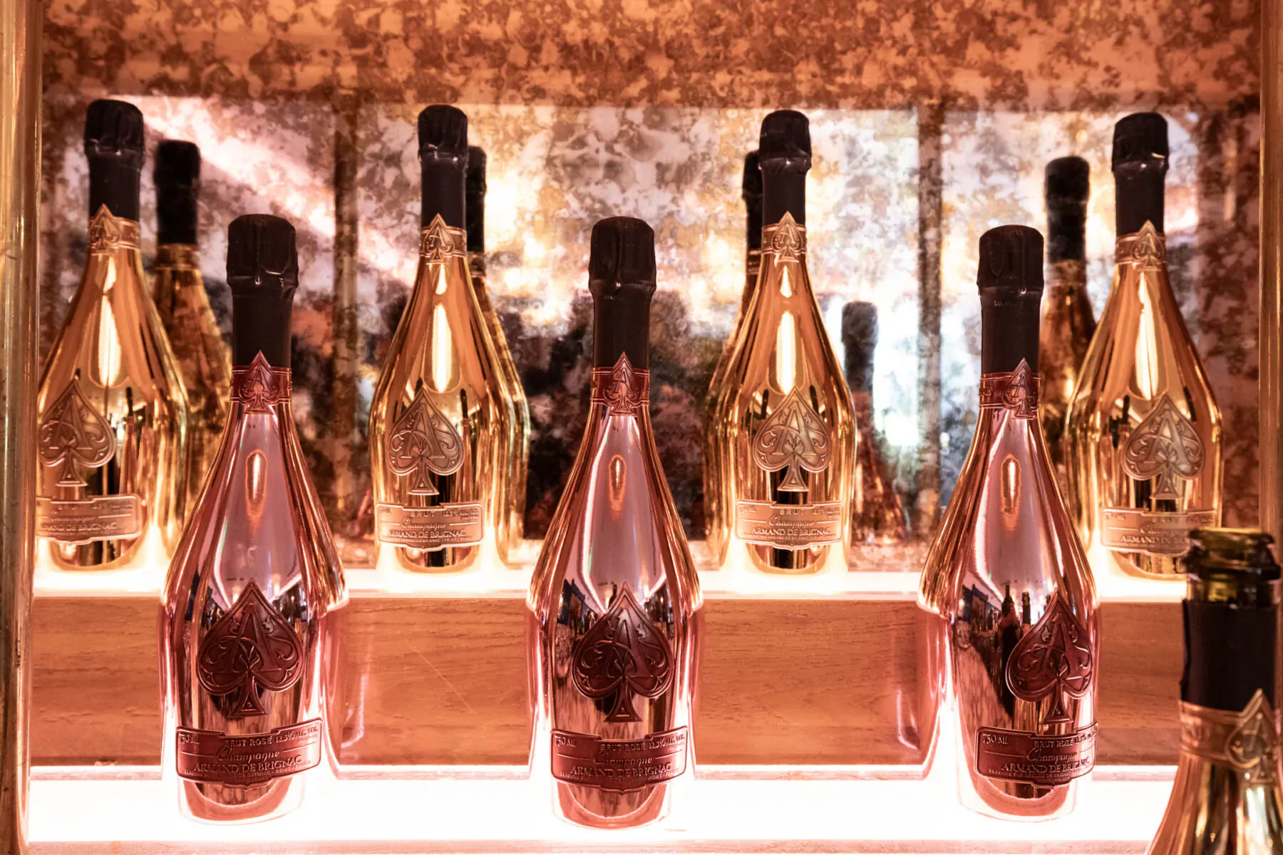 Row of glowing gold and pink Armand de Brignac champagne bottles on illuminated wooden shelf with mirrored backdrop