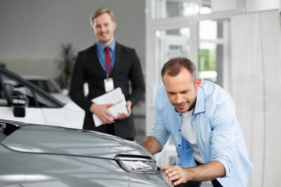 Salesman in suit holding papers stands behind customer in blue shirt inspecting gray used car's headlight in showroom