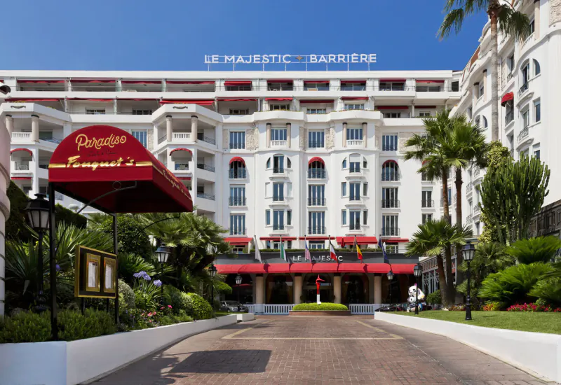 Le Majestic Barrière luxury hotel in Cannes with palm trees, red awnings, and grand entrance; 'Palais' sign visible.