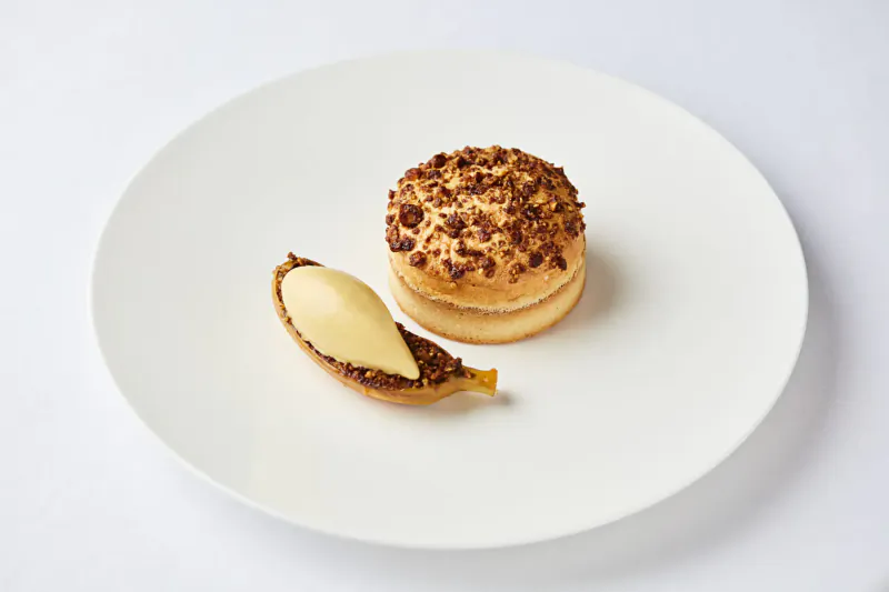 White plate with crumb-topped pastry round and quenelle of custard on caramelized banana leaf