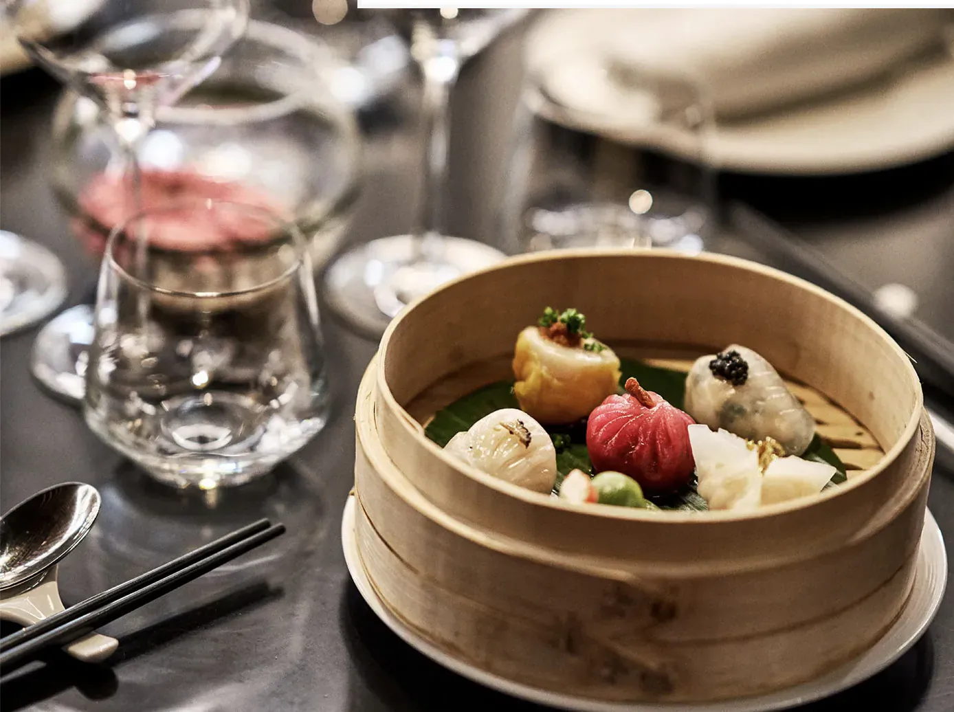 Assorted colourful steamed dumplings in bamboo steamer on elegant table with wine glasses at Chai Wu Harrods