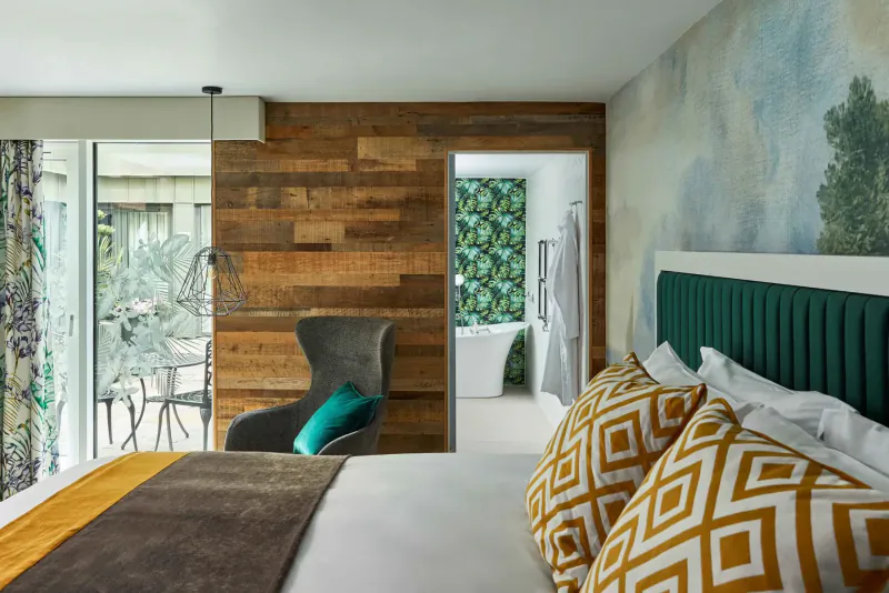 Cozy Hotel Indigo Bath bedroom with wood accent wall, green headboard bed with yellow pillows, turquoise armchair, and lush mural.