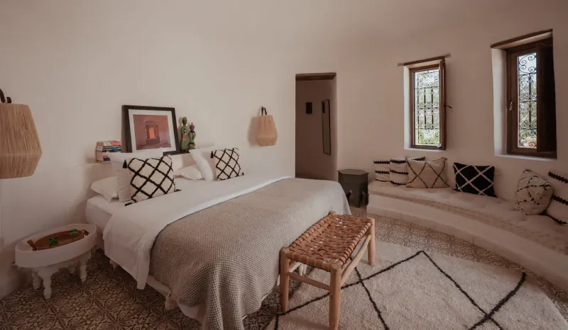 Cozy Moroccan bedroom in curved white room with king bed, woven pillows, bench, plants, and rattan lamps at Les Jardins De Villa Maroc