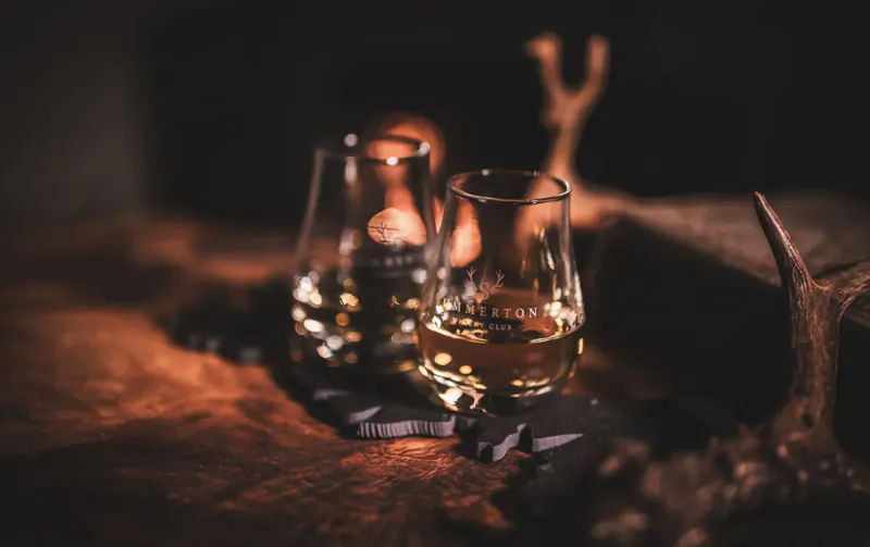 Two Glendronach whisky glasses with amber liquid on fur mat beside antlers in warm-lit rustic setting