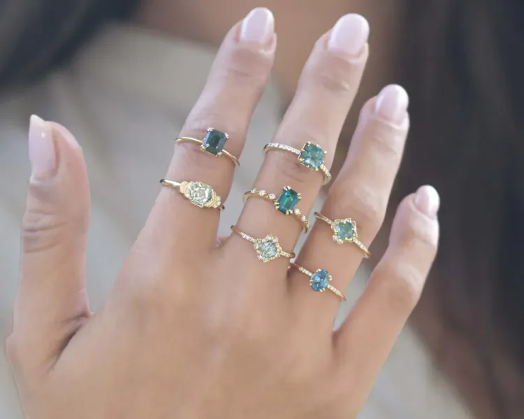 Woman's hand displaying multiple gold rings with green gemstones, various styles stacked on fingers.