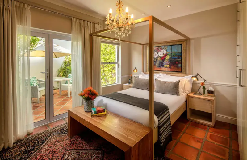 Elegant bedroom at Spanish Farm Guest Lodge with four-poster bed, chandelier, artwork, flowers, and terrace view.