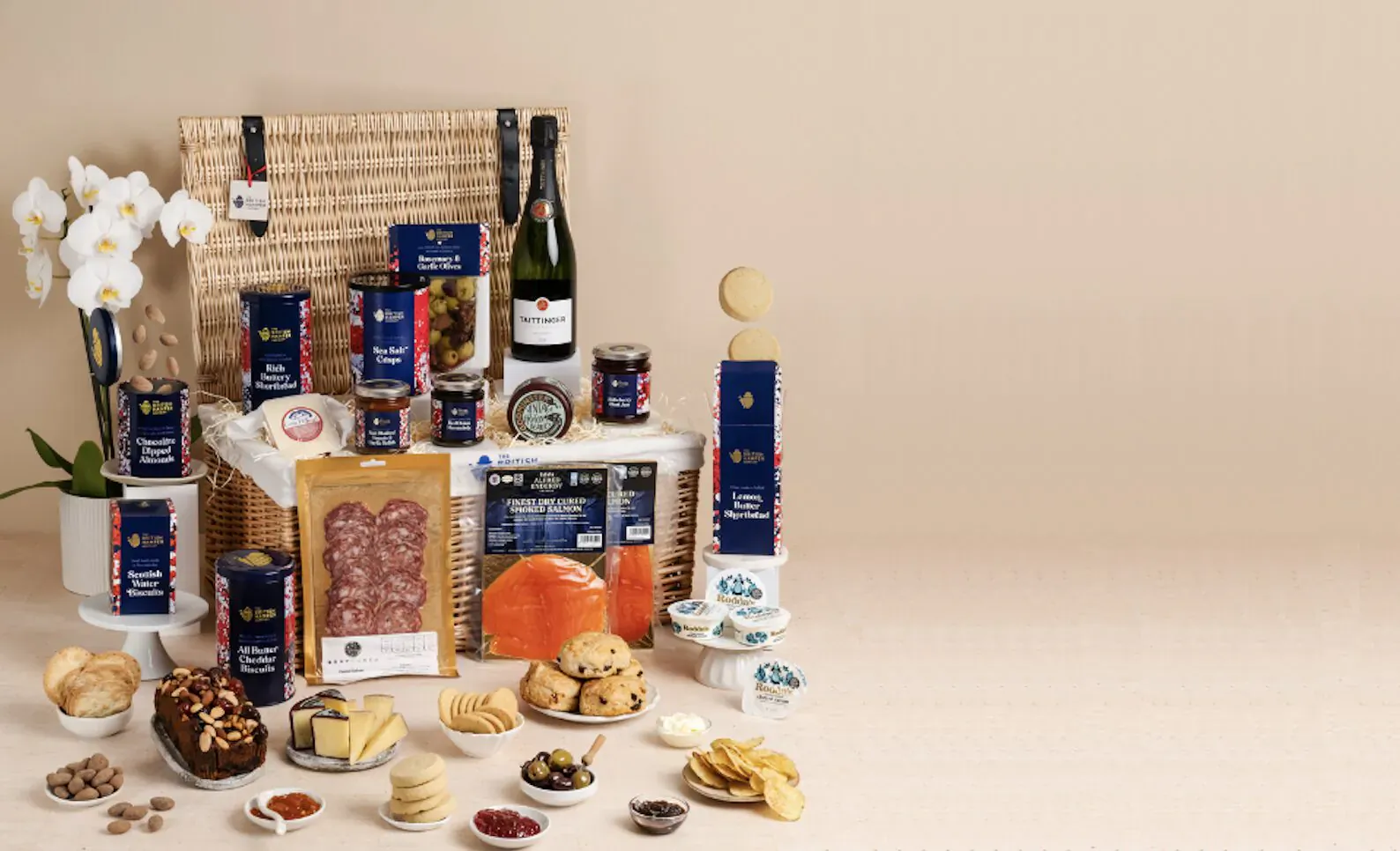 Wicker hamper basket overflowing with champagne, cheeses, salami, biscuits, and gourmet treats on beige backdrop with orchids.