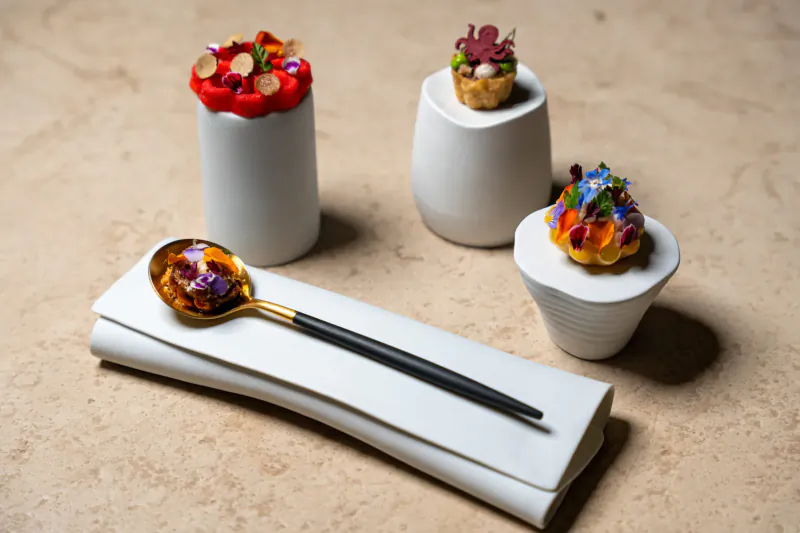 Assortment of elegant gourmet dishes in white ceramic cups topped with colorful flowers and nuts, gold spoon on napkin