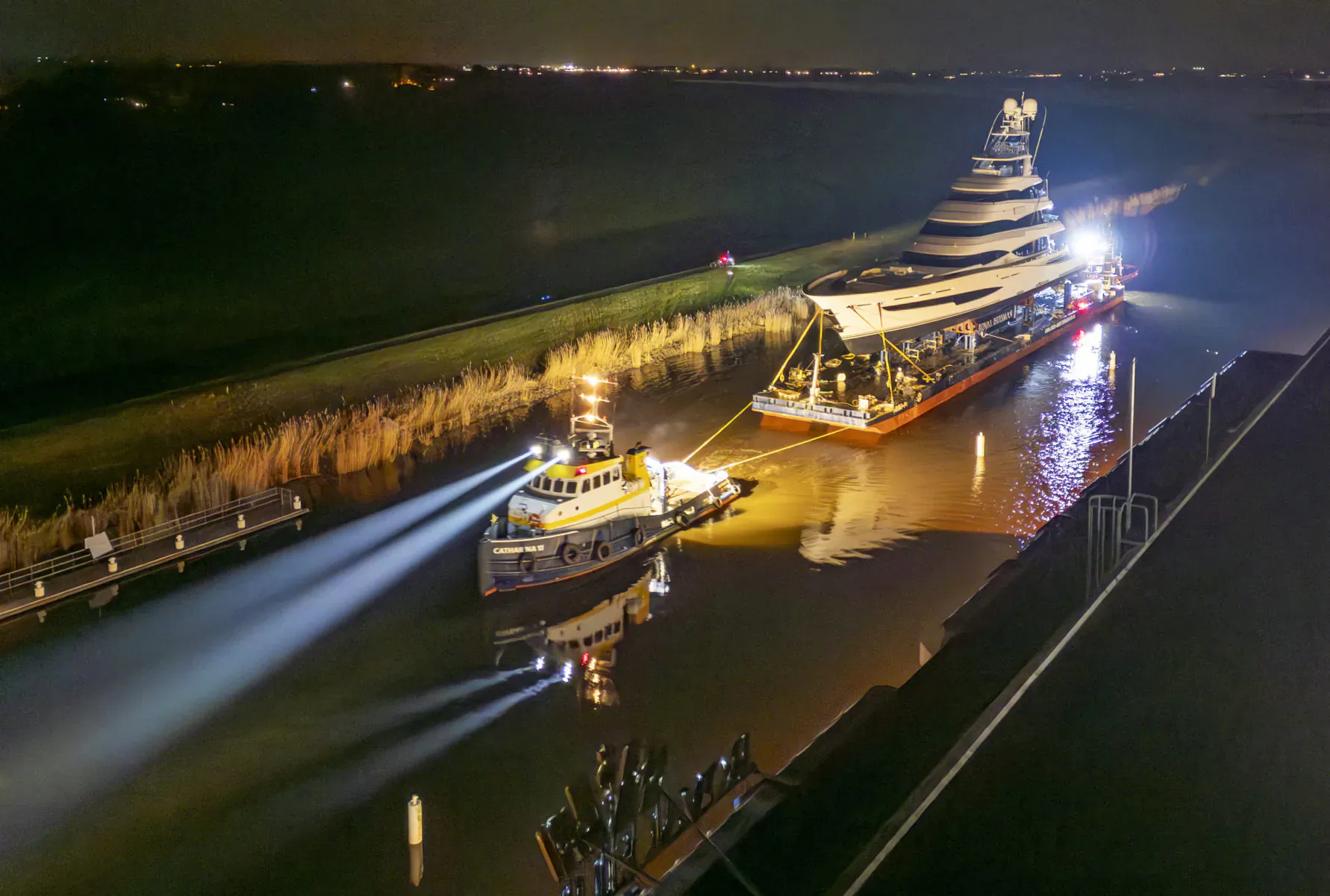 Night scene of illuminated multi-tiered barge towed by tugboat on canal, lights reflecting on water.