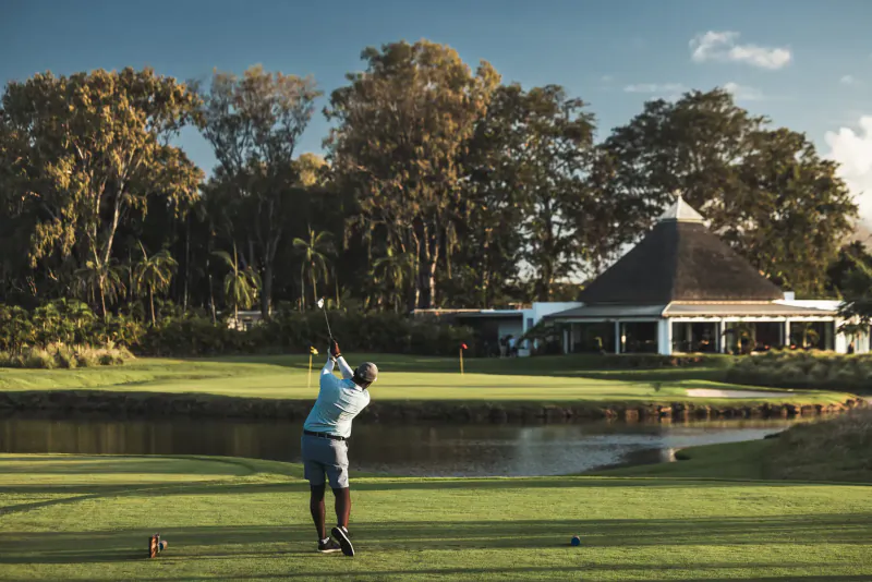 Golfer mid-swing on lush green near pond, thatched clubhouse, and palm trees at Mauritius golf course