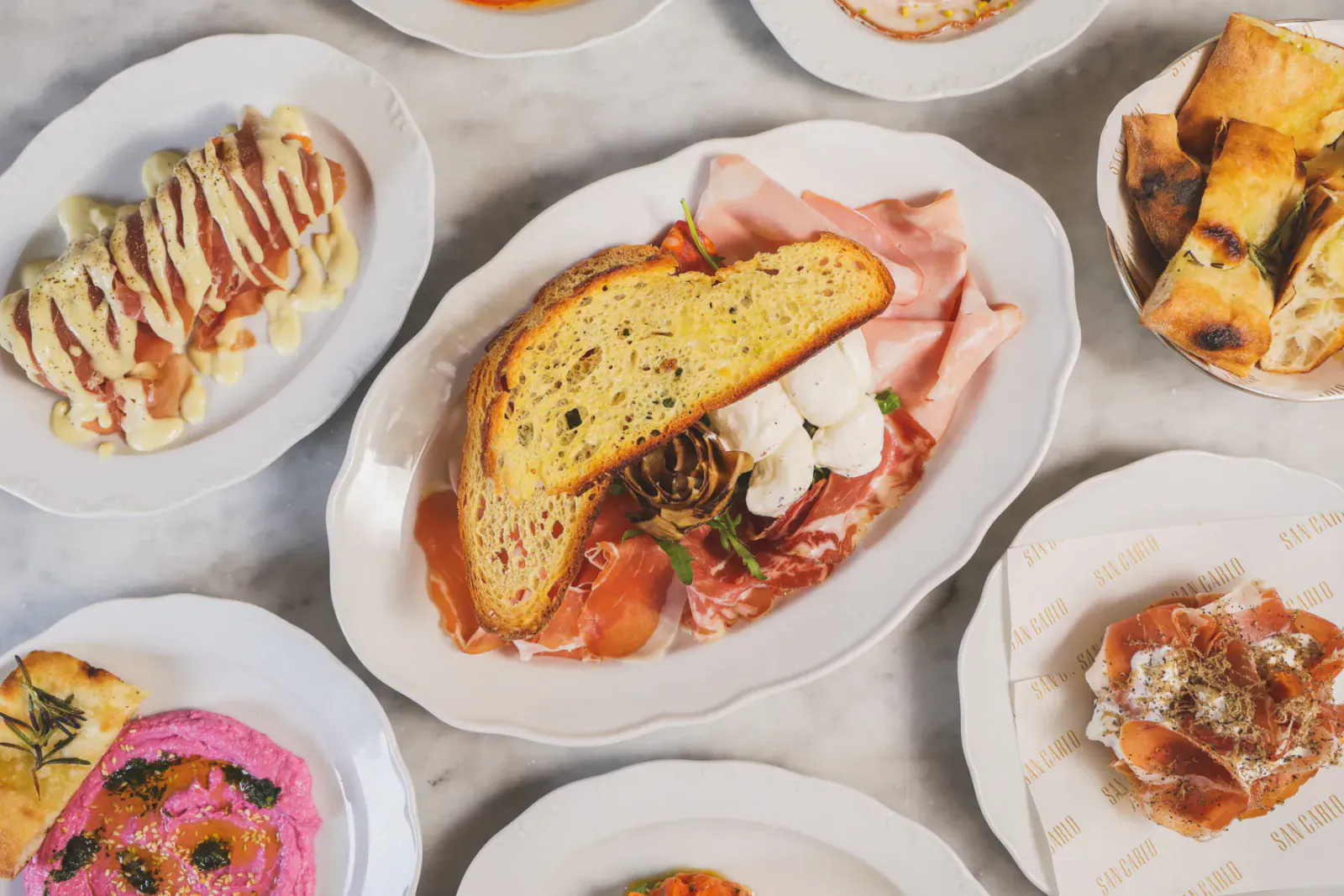 Assortment of Italian dishes on white plates: prosciutto sandwiches, salmon with sauce, pink hummus, bread and focaccia