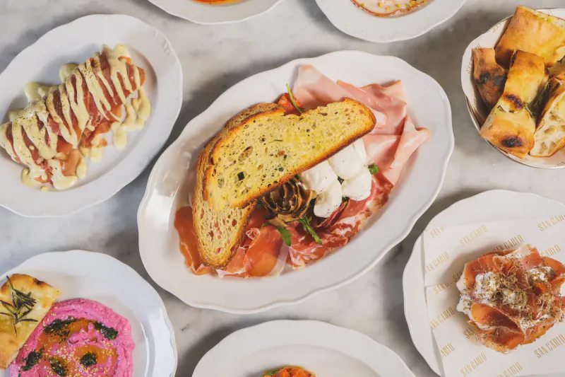 Assortment of Italian dishes on white plates: prosciutto sandwiches, salmon with sauce, pink hummus, bread and focaccia