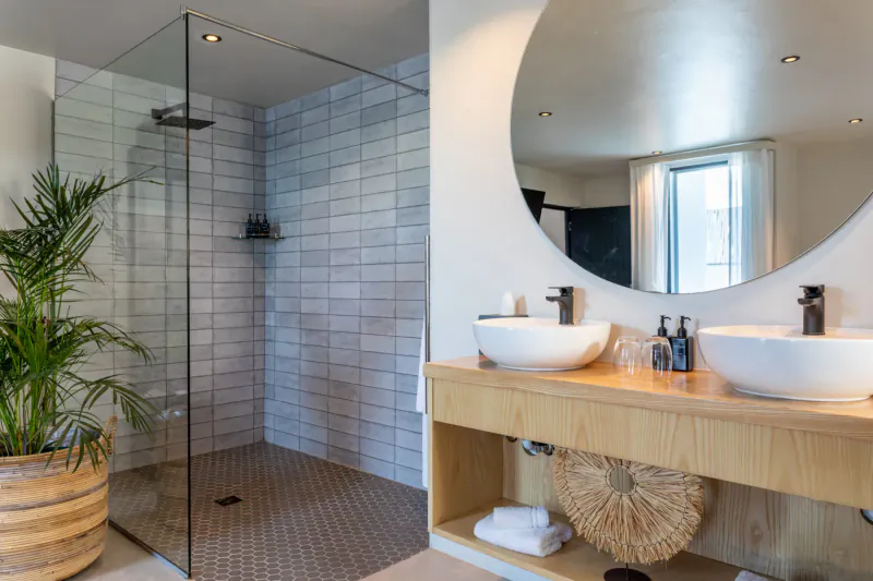 Modern hotel bathroom with glass shower, wood vanity, two sinks, round mirror, plants, and towels in Oyster Suite.