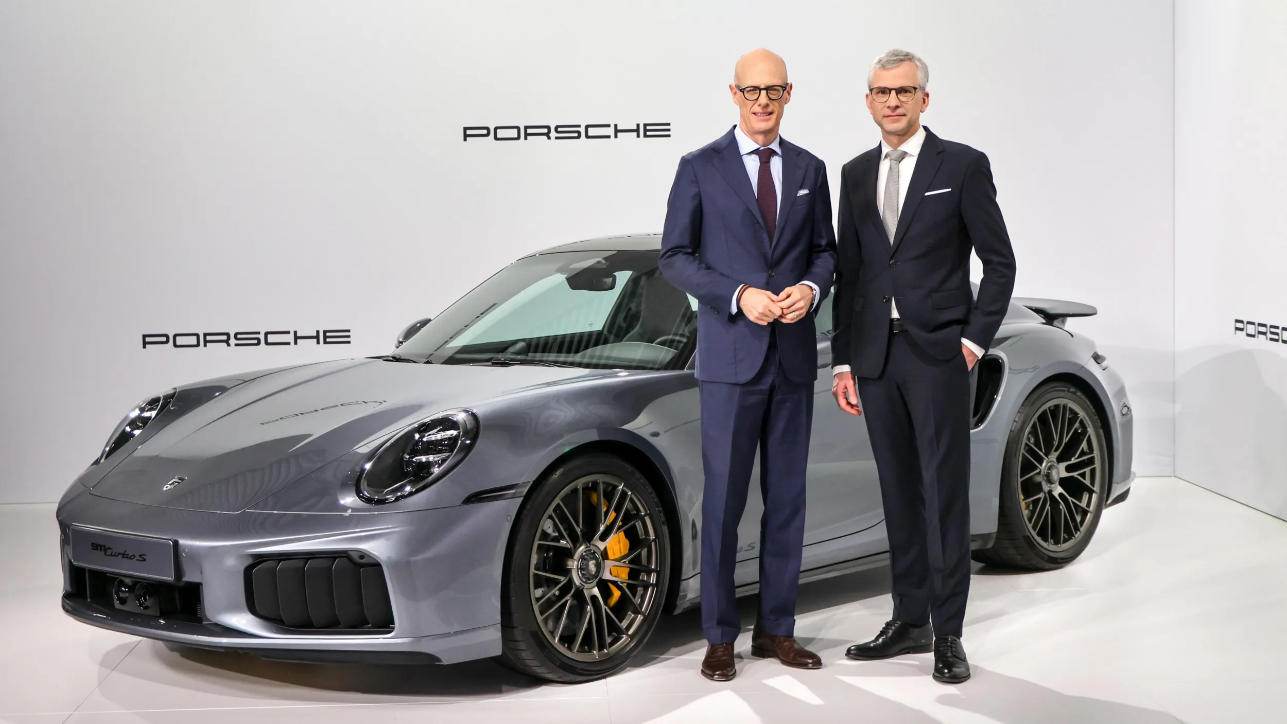 Two suited executives, one with glasses, stand beside a sleek gray Porsche 911 sports car in a studio.