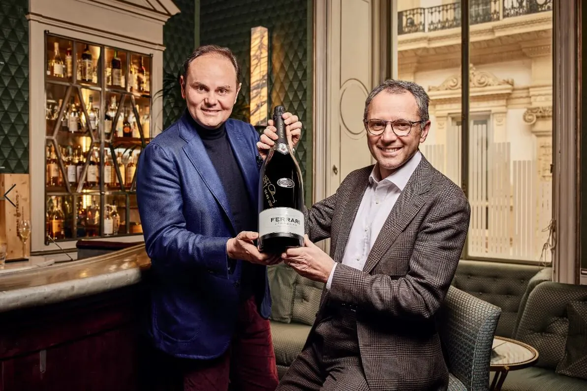 Matteo Lunelli and colleague holding Ferrari Trento bottle in elegant bar with liquor shelves.