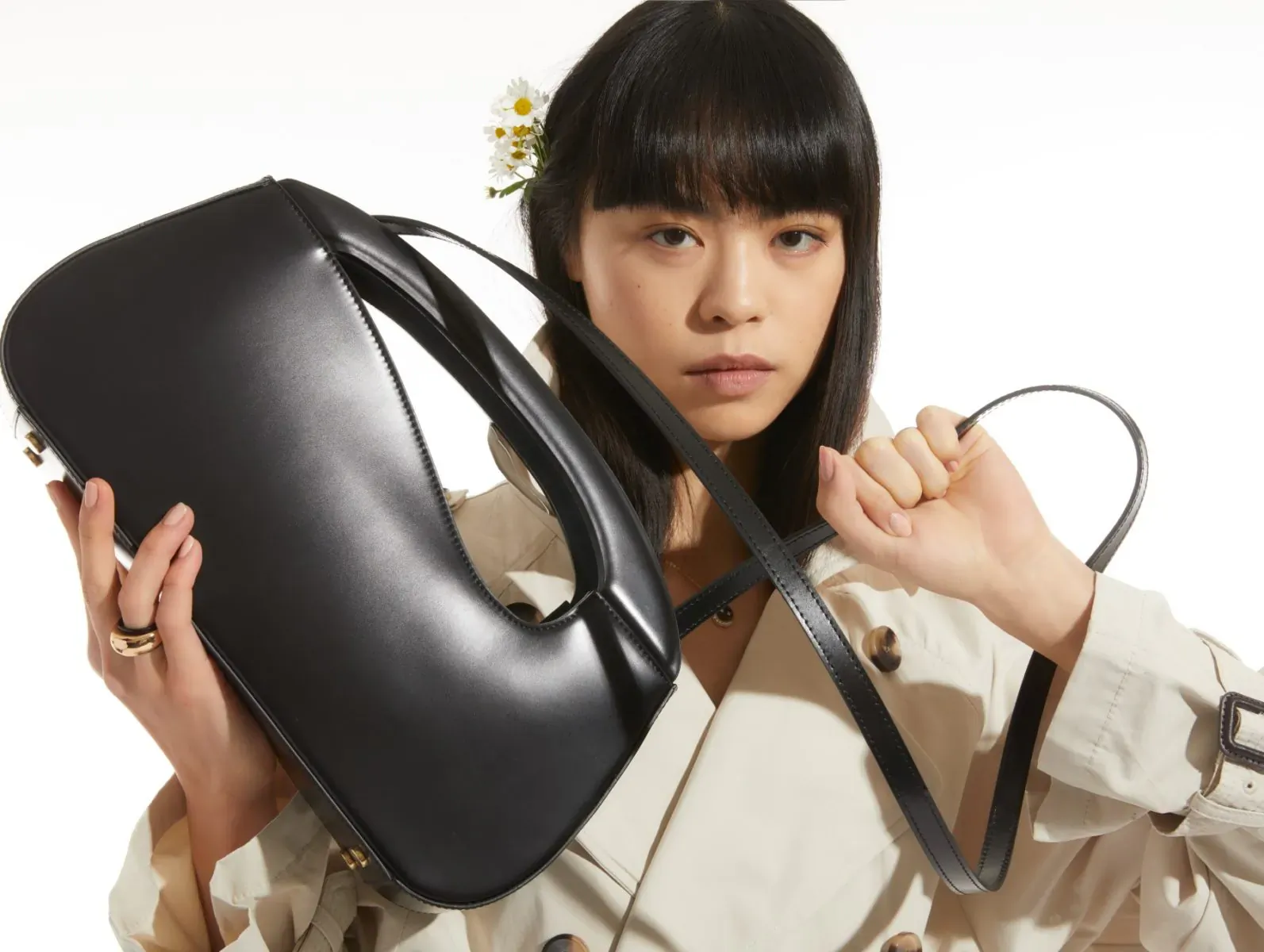 Asian woman with daisy in black bangs holds black leather hobo bag, wearing beige trench coat